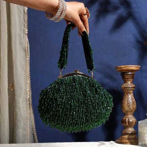 Könnte beinhalten: Eine grüne, mit Perlen besetzte Handtasche mit goldenem Verschluss und Griff. Die Tasche ist mit langen, herabhängenden Perlen bedeckt, die einen Fransen-Effekt erzeugen. Der Griff ist aus dunkelgrünem Samt. Die Tasche wird von einer Hand mit silbernen Armbändern und Ringen gehalten.