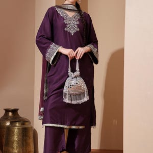 Könnte beinhalten: Ein pflaumenfarbenes Salwar-Kameez-Ensemble mit aufwendiger silberner Stickerei an Ausschnitt, Ärmeln und Dupatta. Das Outfit umfasst ein passendes Oberteil, weite Hosen und einen Schal. Eine kleine, silberne Perlenbeutel wird in der Hand gehalten.