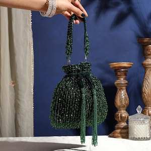 Peut inclure: Un sac à main à cordon de serrage vert foncé recouvert de perles de verre complexes. Le sac présente une anse perlée assortie et deux longs pompons perlés pendants suspendus au cordon central, créant un aspect texturé et scintillant.