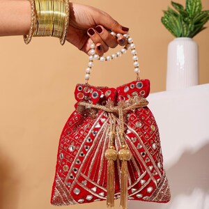 Può includere: Una borsa rossa con coulisse con ricami dorati e specchi. La borsa presenta un manico di perle e nappe dorate. Una pila di braccialetti d'oro adorna il polso che tiene la borsa. Un accessorio festivo.
