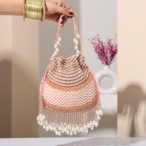 May include: A beaded drawstring bag in shades of tan, pink, and white. The bag features a layered design with rows of beads and a fringe of dangling pearls. The handles are adorned with pearls, and the bag is held by a hand wearing gold bangles.