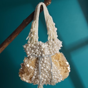 Puede incluir: Un pequeño bolso de mano con cuentas de color crema con un asa de perlas y un cierre de cordón. El bolso está adornado con perlas y lentejuelas, y cuelga de una rama de madera sobre un fondo turquesa.