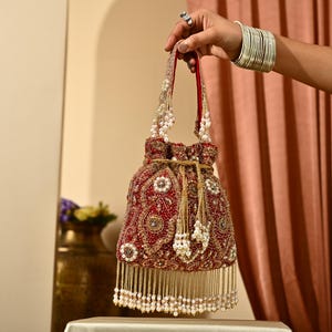 Może przedstawiać: Czerwona torebka ze sznurkiem, z koralikami i złotymi zdobieniami. Torebka ma frędzle i paski ozdobione perłami. Jest trzymana przez dłoń z srebrnymi bransoletkami i pierścionkiem. Modny dodatek.