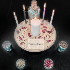 May include: A circular wooden tray holds a central white pillar candle, four smaller taper candles in white, black, and purple, a glass bottle of teal-tipped matches, small jars, and dried rose petals. A card reads your spell here.