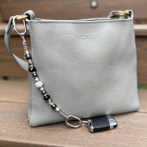 May include: A gray leather crossbody bag with a beaded strap and a key fob. The bag has a simple, square design with a top handle and a silver-tone clasp. The beaded strap features black, silver, and clear beads. The bag is sitting on a wooden surface.