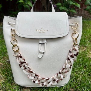 May include: White leather backpack with a gold chain strap and a brown and white plastic chain strap. The bag has a gold buckle and the text "kate spade new york" is printed on the front.