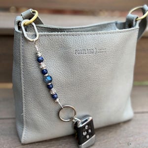 May include: A gray leather handbag with a beaded keychain attached. The keychain features blue beads and a silver ring holding a car key fob. The bag has gold-colored hardware and the text "PORTLAND LEATHER GOODS" is visible.