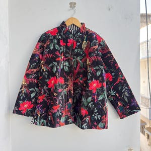 Könnte beinhalten: Schwarze Steppjacke mit einem lebendigen Blumenmuster aus roten Blüten und grünen Blättern. Die Jacke hat einen Stehkragen und lange Ärmel und hängt an einem Holzbügel vor einer weißen Wand.