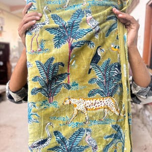 Puede incluir: Un rollo de tela con un diseño de jungla. La tela tiene un fondo amarillo-verdoso con ilustraciones de leopardos, tucanes, flamencos y palmeras en tonos de azul, blanco y marrón.