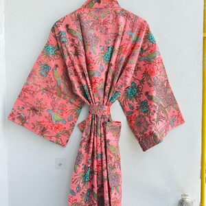 May include: A pink kimono robe featuring a repeating pattern of colourful birds, including green and yellow pheasants, interspersed with teal and red floral motifs. The garment has wide sleeves, a matching tie belt, and a wrap-front design.