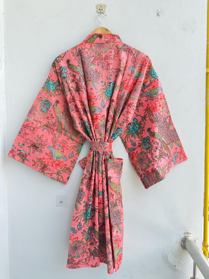 May include: A pink kimono robe featuring a repeating pattern of colourful birds, including green and yellow pheasants, interspersed with teal and red floral motifs. The garment has wide sleeves, a matching tie belt, and a wrap-front design.
