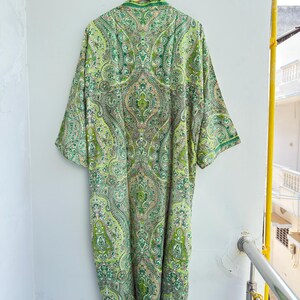 May include: A long-sleeved robe featuring an intricate paisley pattern in various shades of green, lime, and cream. The garment has a mandarin collar, a straight silhouette, and is displayed hanging on a wooden hanger against a plain wall.