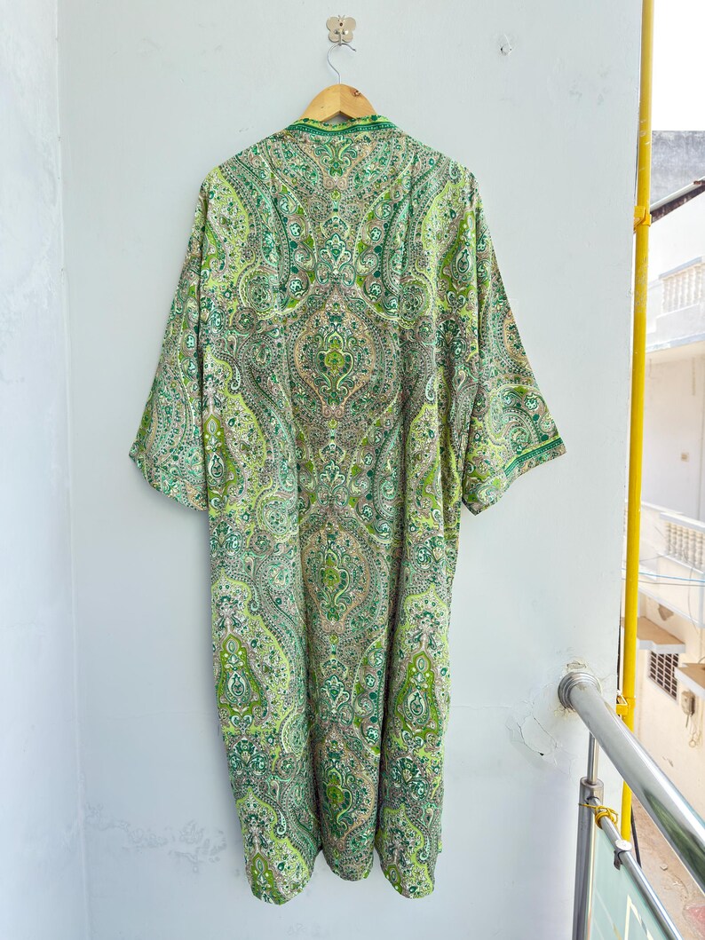 May include: A long-sleeved robe featuring an intricate paisley pattern in various shades of green, lime, and cream. The garment has a mandarin collar, a straight silhouette, and is displayed hanging on a wooden hanger against a plain wall.
