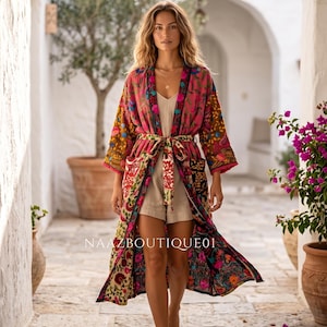 May include: A long, colorful kimono with a floral pattern, worn with a matching belt. The kimono features shades of pink, yellow, and green. The text "NAAZBOUTIQUE01" is visible on the garment. The model is walking on a stone path.