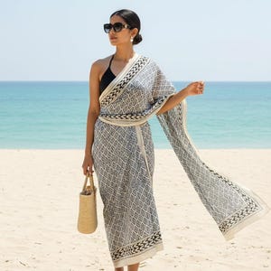 Könnte beinhalten: Eine Frau am Strand mit einem schwarzen Badeanzug und einem gemusterten Wickel. Der Wickel hat ein geometrisches Design in Schwarz und Weiß. Sie hält auch eine gewebte Tragetasche und trägt eine Sonnenbrille.