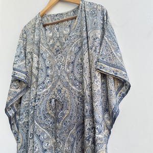 May include: A kaftan featuring a detailed blue, grey, and beige paisley pattern throughout the fabric. The garment has a V-neckline, wide short sleeves, and a drawstring waist detail, hanging from a wooden hanger against a plain white wall.