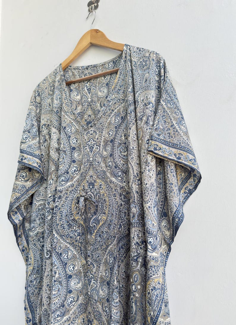 May include: A kaftan featuring a detailed blue, grey, and beige paisley pattern throughout the fabric. The garment has a V-neckline, wide short sleeves, and a drawstring waist detail, hanging from a wooden hanger against a plain white wall.