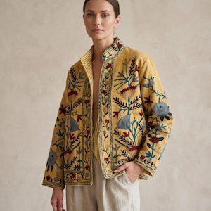 以下が含まれることがあります： 赤、青、緑の花柄刺繍が施されたマスタードイエローのジャケット。 ジャケットはマンダリンカラーで、裏地はストライプ柄。 袖は長く、ジャケットはフロントオープンです。