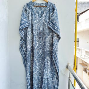 May include: A floor-length kaftan dress featuring a blue and white intricate paisley pattern. The garment has a V-neckline, wide short sleeves, and a drawstring waist detail. It is displayed on a wooden hanger against a plain wall.