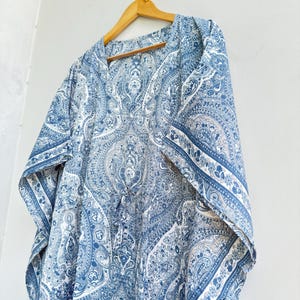 May include: A blue and white kaftan featuring an intricate paisley pattern throughout the fabric. The garment has a V-neckline, wide short sleeves, and a drawstring detail at the centre front, all displayed on a wooden hanger against a wall.