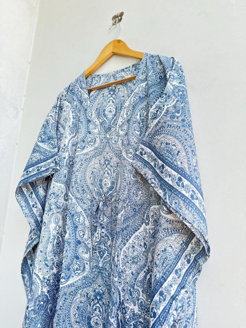 May include: A blue and white kaftan featuring an intricate paisley pattern throughout the fabric. The garment has a V-neckline, wide short sleeves, and a drawstring detail at the centre front, all displayed on a wooden hanger against a wall.
