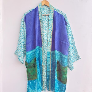May include: A colorful kimono-style robe with a mix of patterns and colors. The robe features a vibrant blue central panel, teal floral sleeves, and turquoise and green accents. It has two pockets and is hanging on a wooden hanger.