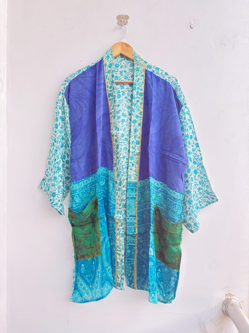 May include: A colorful kimono-style robe with a mix of patterns and colors. The robe features a vibrant blue central panel, teal floral sleeves, and turquoise and green accents. It has two pockets and is hanging on a wooden hanger.