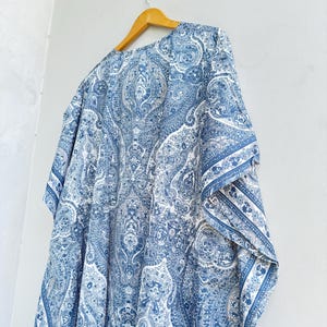May include: A blue and white kaftan featuring an intricate paisley pattern throughout the fabric. The garment has a loose, flowing silhouette with wide sleeves and is displayed on a wooden hanger against a plain, light-coloured wall.