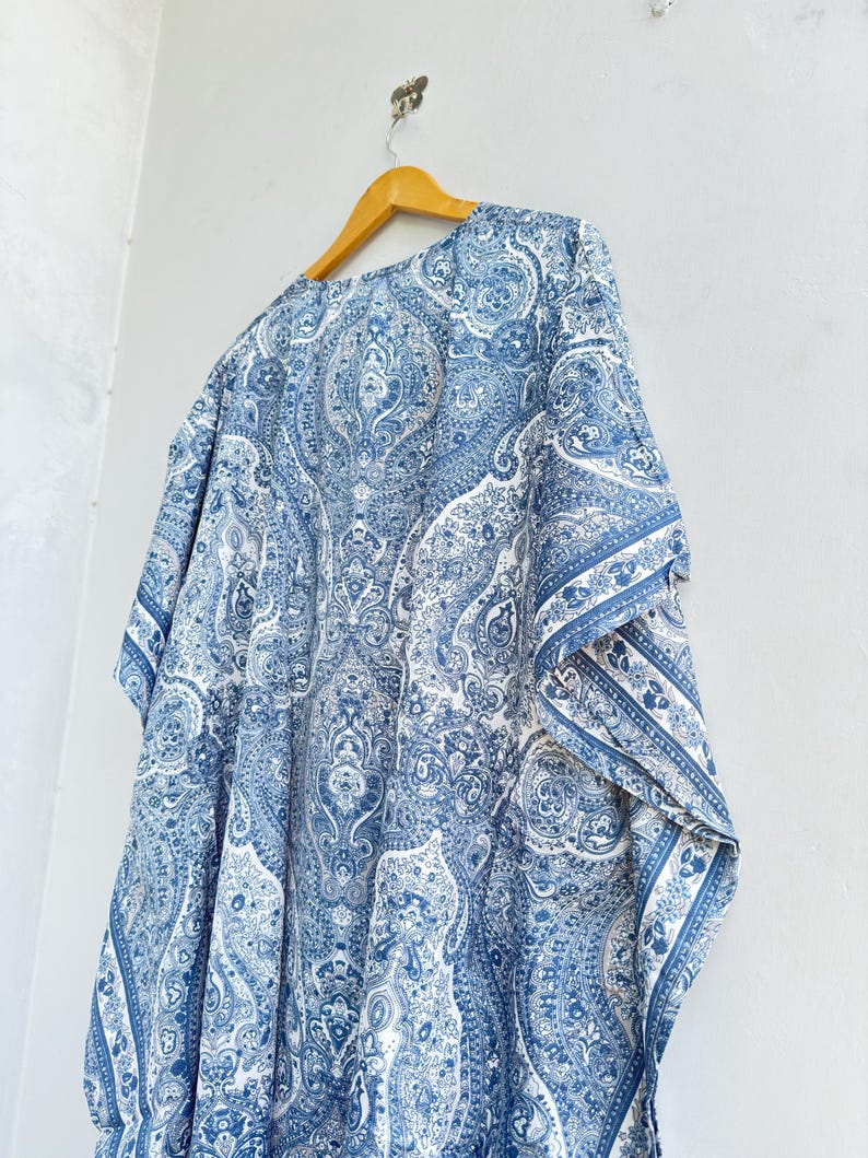 May include: A blue and white kaftan featuring an intricate paisley pattern throughout the fabric. The garment has a loose, flowing silhouette with wide sleeves and is displayed on a wooden hanger against a plain, light-coloured wall.
