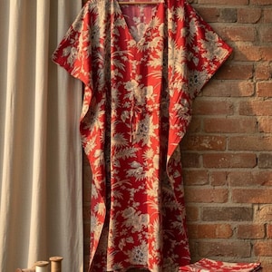 Op de afbeelding: Een rode kaftan-jurk met een crème-kleurige bloemen- en botanische print, een V-hals en een trekkoord in de taille. Het kledingstuk hangt aan een houten kledinghanger en heeft wijde mouwen en een losvallend, vloeiend silhouet tot onder de knie.