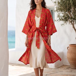 Könnte beinhalten: Roter Kimono-Stil-Bademantel mit sich wiederholendem goldenem Tierdruck, an der Taille mit einem passenden Gürtel gebunden. Der Bademantel wird über einem cremefarbenen Slip-Kleid getragen. Das Bild wird im Freien aufgenommen.