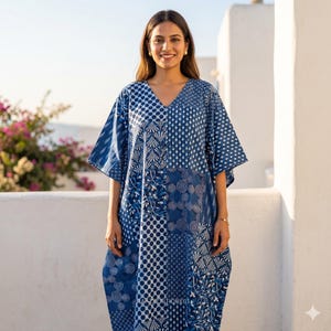 Może przedstawiać: Niebiesko-biała wzorzysta sukienka kaftan z dekoltem w serek. Sukienka ma patchworkowy wzór z różnymi geometrycznymi i kwiatowymi wzorami. Rękawy są krótkie i szerokie. Sukienka jest noszona przez osobę stojącą na zewnątrz w świetle słonecznym.