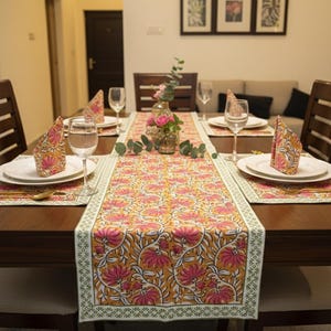 Könnte beinhalten: Ein gedeckter Esstisch mit einem Tischläufer mit Blumenmuster und passenden Platzsets. Auf dem Tisch sind weiße Teller, gefaltete Servietten und Weingläser angeordnet. In der Mitte steht eine Vase mit Blumen.