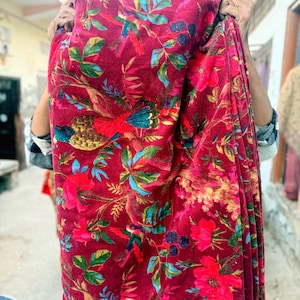 May include: A deep burgundy fabric with a colourful floral and bird pattern. The design includes red, blue, green, and yellow details. The fabric is held up, displaying the intricate print and rich colour palette.