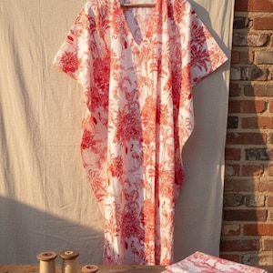 Può includere: Un caftano bianco e rosso fantasia con scollo a V, appeso a una gruccia di legno. Il tessuto presenta un design tropicale con fenicotteri e palme. Una pila di tessuto abbinato e materiale da cucito sono su un tavolo di legno.