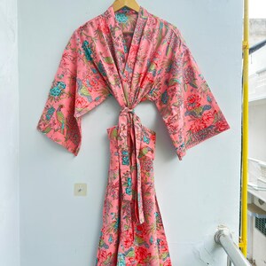 May include: A pink kimono-style robe featuring a vibrant floral and bird pattern in shades of teal, red, and green. The garment has wide, three-quarter length sleeves, a V-neckline, and a matching fabric tie belt at the waist for closure.