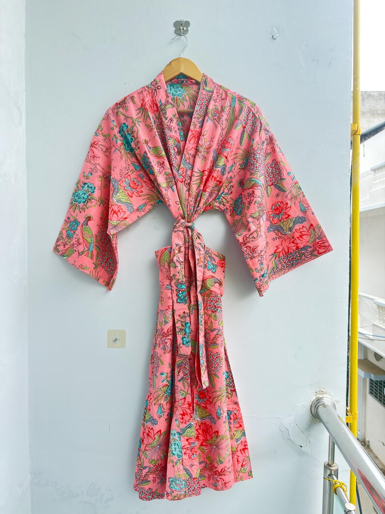 May include: A pink kimono-style robe featuring a vibrant floral and bird pattern in shades of teal, red, and green. The garment has wide, three-quarter length sleeves, a V-neckline, and a matching fabric tie belt at the waist for closure.