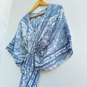 May include: A blue and white paisley print top featuring a V-neckline, wide kimono-style sleeves, and a front tie closure. The garment is displayed on a wooden hanger, showcasing a detailed intricate pattern throughout the fabric design.