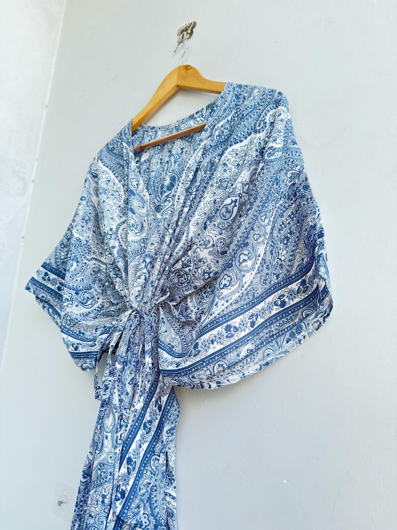 May include: A blue and white paisley print top featuring a V-neckline, wide kimono-style sleeves, and a front tie closure. The garment is displayed on a wooden hanger, showcasing a detailed intricate pattern throughout the fabric design.