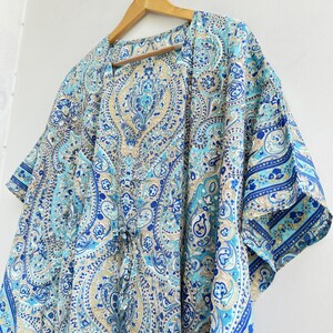 May include: A kaftan featuring a detailed paisley pattern in shades of blue, turquoise, and beige. The garment has short, wide sleeves, a V-neckline, and a drawstring waist tie. The fabric displays intricate swirling motifs and floral elements.