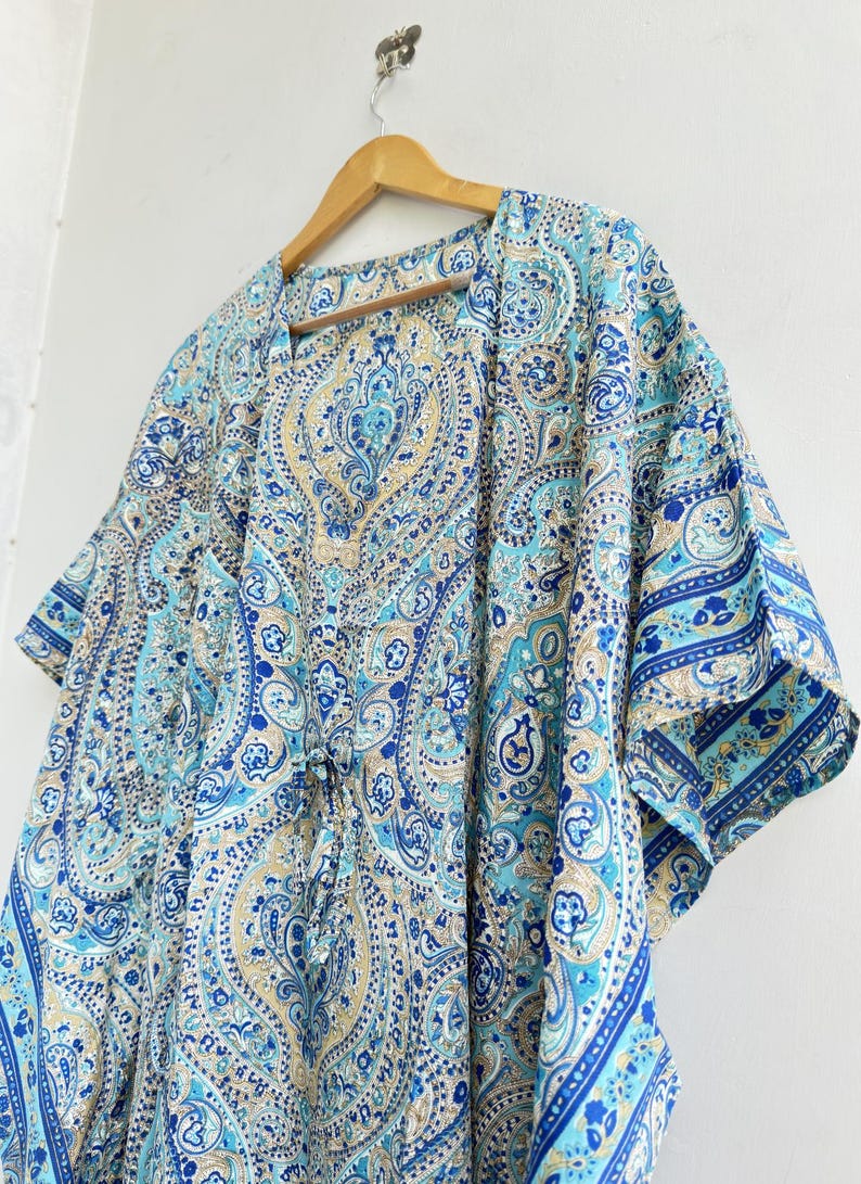 May include: A kaftan featuring a detailed paisley pattern in shades of blue, turquoise, and beige. The garment has short, wide sleeves, a V-neckline, and a drawstring waist tie. The fabric displays intricate swirling motifs and floral elements.