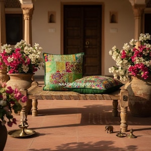 Op de afbeelding: Een buitenscène met een bank met twee patchwork kussens. De kussens hebben groene, rode en gele patronen. Grote vazen met roze en witte bloemen staan aan weerszijden. De tekst "NAAZBOUTIQUE01" is zichtbaar op een kussen.