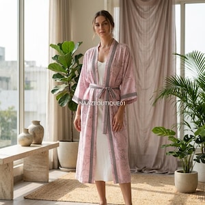 Könnte beinhalten: Ein hellrosa Kimono-ähnlicher Bademantel mit floralem Muster und passendem Gürtel. Der Bademantel hat schwarz-weiße Verzierungen und wird über einem weißen Kleid getragen. Der Text "NAAZBOUTIQUE01" ist sichtbar.