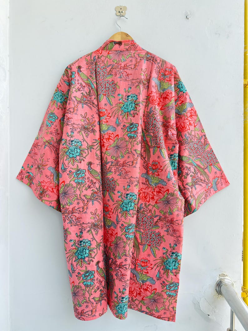 May include: A long-sleeved kimono robe in a vibrant coral pink hue, featuring an intricate all-over print of teal blue flowers, green foliage, and small birds perched on branches. The garment has a wide, open front design and loose sleeves.