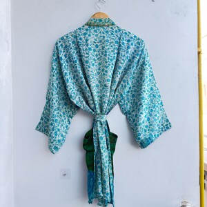 May include: A light blue floral print kimono robe hanging on a wooden hanger. The robe is tied at the waist with a matching sash. The robe has a green and blue accent on the bottom.