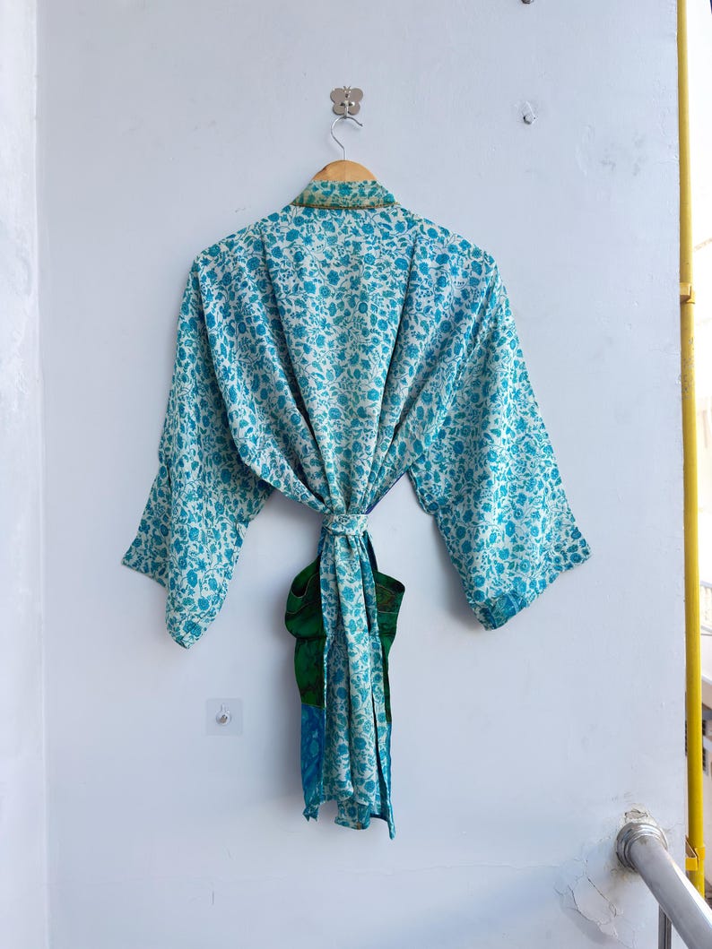 May include: A light blue floral print kimono robe hanging on a wooden hanger. The robe is tied at the waist with a matching sash. The robe has a green and blue accent on the bottom.