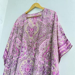 May include: A loose-fitting kaftan featuring a vibrant pink, purple, and light green paisley pattern. The garment has a V-neckline, short wide sleeves, and a flowing silhouette, displayed on a wooden hanger against a plain light background.