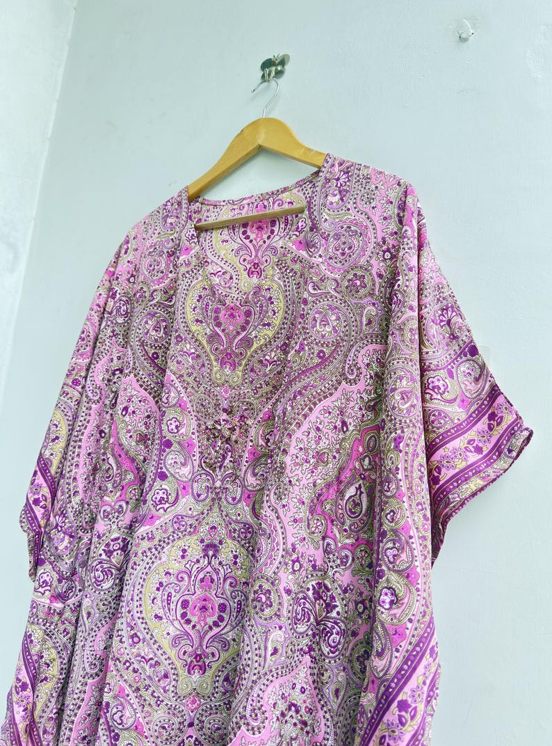 May include: A loose-fitting kaftan featuring a vibrant pink, purple, and light green paisley pattern. The garment has a V-neckline, short wide sleeves, and a flowing silhouette, displayed on a wooden hanger against a plain light background.
