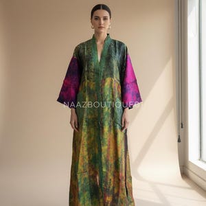 Pode incluir: Robe comprido, aberto na frente, com um padrão abstrato vibrante. O robe apresenta decote em V e mangas compridas com punhos magenta. O corpo do robe é uma mistura de tons verdes, amarelos e azuis. O texto "NAAZBOUTIQUE01" é visível.