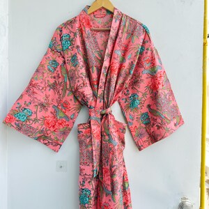 May include: A long-sleeved kimono robe in a vibrant coral pink base colour, featuring an intricate floral and bird pattern in shades of teal, green, and red. The garment includes a matching fabric tie belt at the waist and wide sleeves.